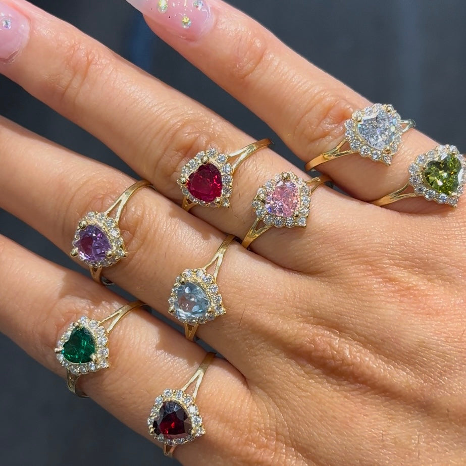 A collection of heart-shaped birthstone rings set in 10k gold with varying colors of gemstones and a halo accent around each.