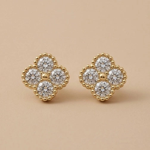 A pair of 10K Gold White Clover Earrings with a four-petal flower design, featuring clear round-cut cubic zirconia surrounded by a beaded gold border, set against a solid beige background.