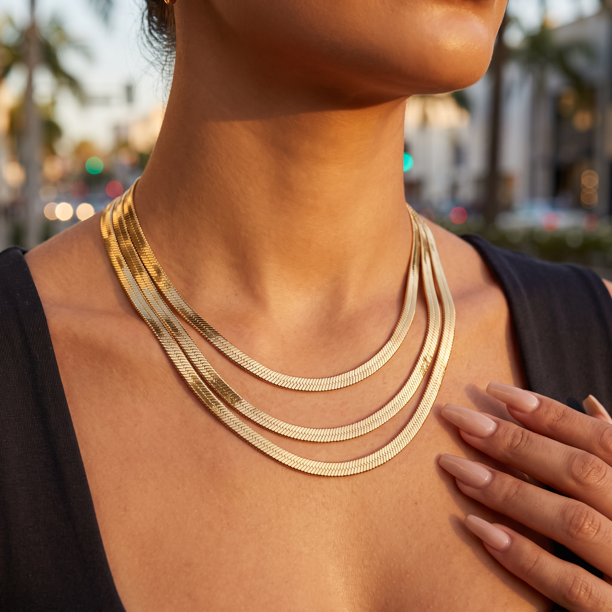 A close-up of a model wearing a layered 10k Gold Herringbone Chain, set against a softly blurred outdoor city background at sunset.