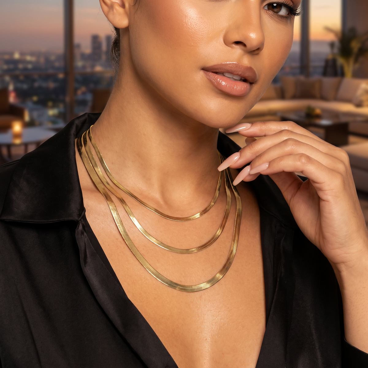 A close-up lifestyle shot of a model wearing a multi-layered 10k Gold Herringbone Chain with a black satin blouse, set against a blurred modern living room background at sunset.
