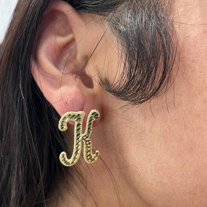 A close-up image of a person wearing 10K gold large cursive initial earrings with a shiny diamond cut finish.